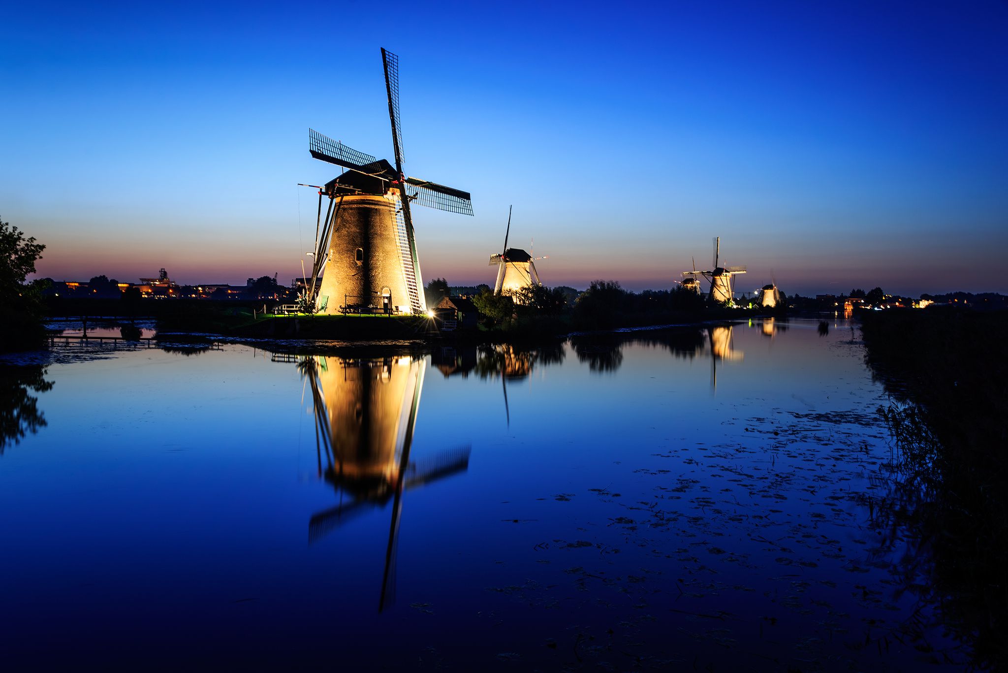 photo of historic illuminated windmills Nederwaard Molen No5 in Dutch Kinderdijk with reflection in the water of the canal after sunset in the beginning of the night in the Netherlands.