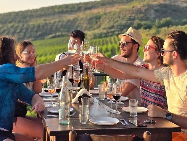 A group of friends toasts with wine glasses at an outdoor table in Durrës, surrounded by green vineyards and hills..jpg
