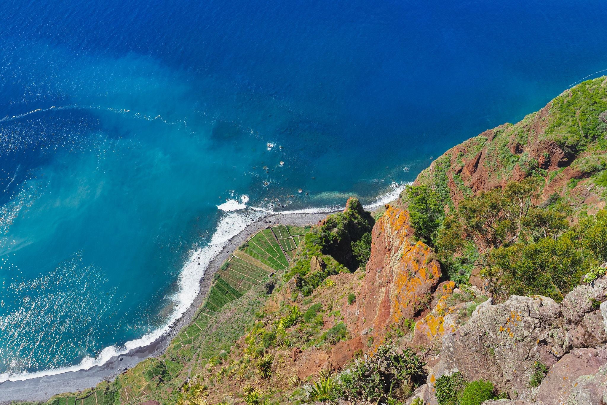 View down from the 'Miradouro do Cabo Girão' viewpoint, Câmara de Lobos. Blue Atlantic ocean and rocky coast with green agricultural fields. Beautiful volcanic landscape of Madeira island, Portugal.