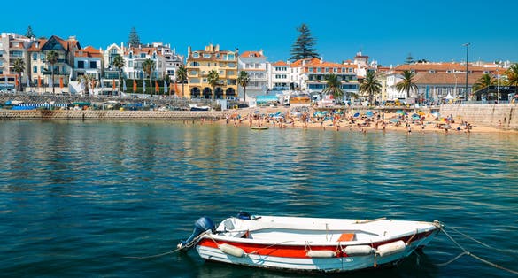 Photo of fishing boat on foreground with crowded sandy beach in Cascais near Lisbon, Portugal during the summer.