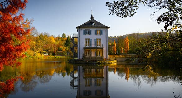 Photo of A baroque house, called Trappenseeschlösschen, surrounded by Lake Trappensee in Heilbronn, Germany during autumn. The house and the colorful trees reflect in the water. 