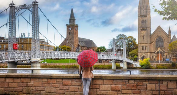 Photo of tourist woman with a scottish pattern umbrella enjoys the view to the cityscape of Inverness, Scotland, during autumn time.