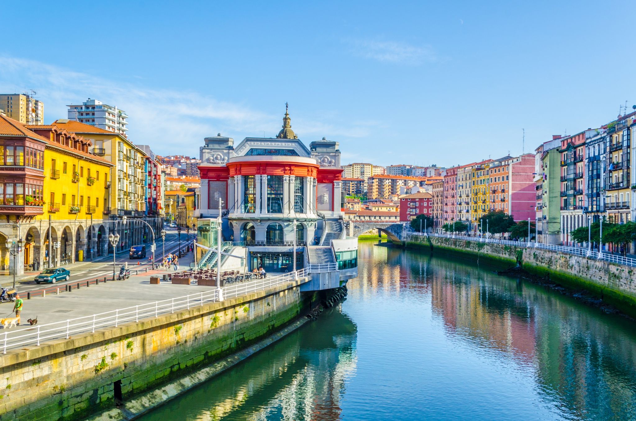 Photo of aerial view of Bilbao, Spain city downtown with a Nevion River, Zubizuri Bridge and promenade. Mountain at the background, with clear blue sky.