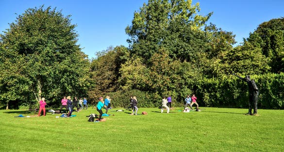 Photo of big group of adult people attending outdoor class, practicing social distancing and exercising yoga outside on lawn on sunny warm autumn day in Marlay Park, Dublin, Ireland.