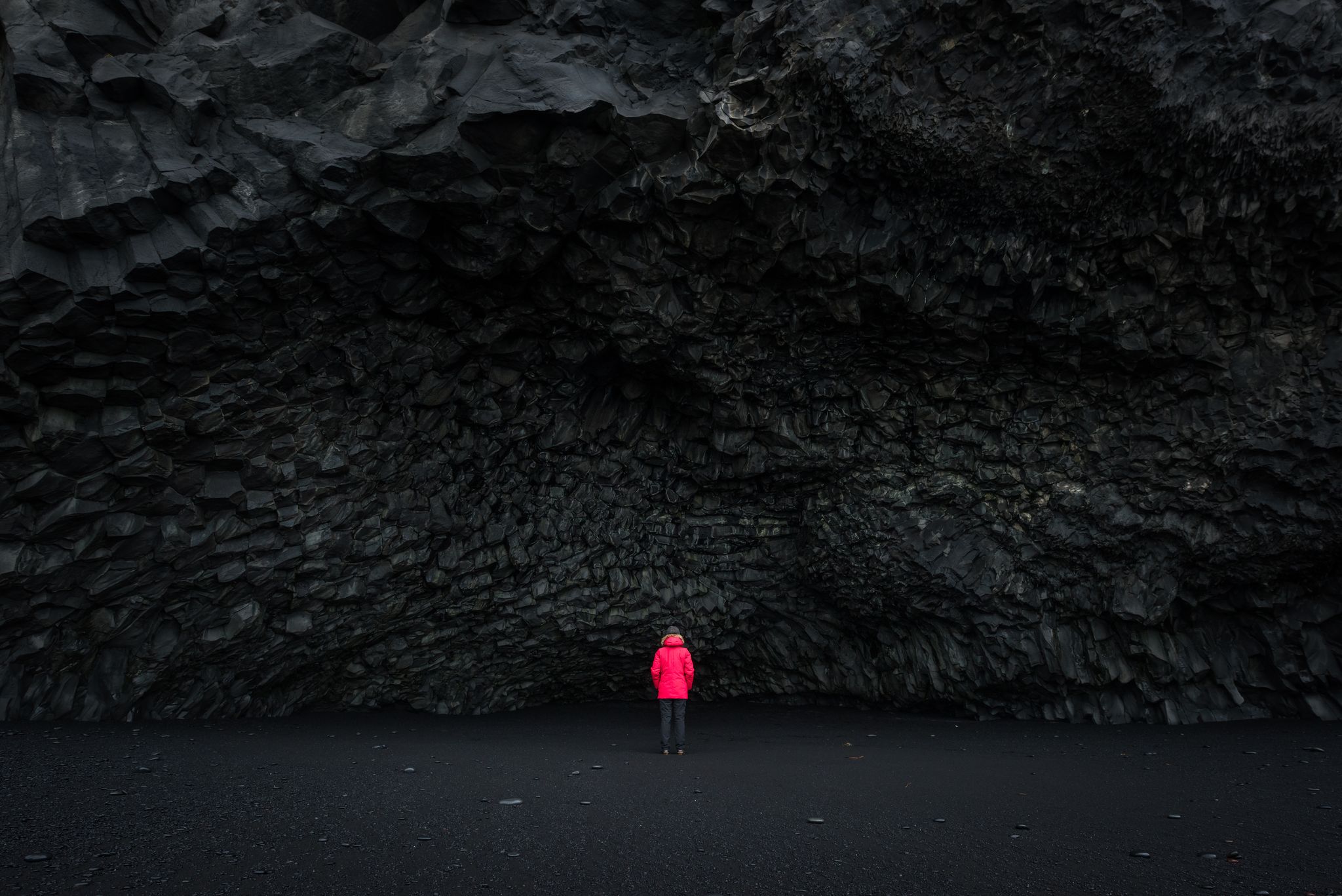 photo of woman in a red coat standing in front of Halsanefshellir Cave on a black sand beach in Iceland.