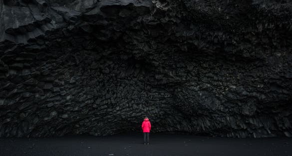 photo of woman in a red coat standing in front of Halsanefshellir Cave on a black sand beach in Iceland.