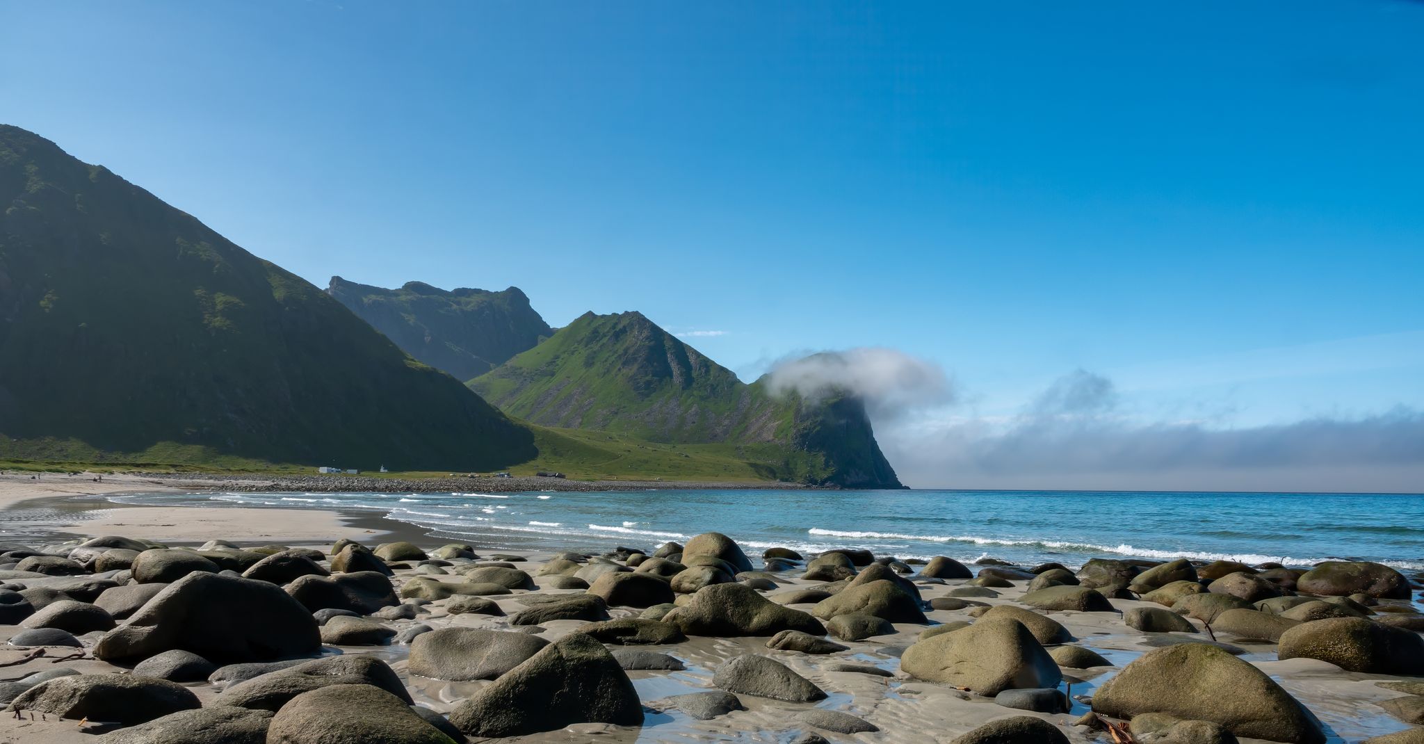 Smooth round stones lie scattered across the sand at Uttakleiv Beach, near Leknes in Lofoten, with turquoise waves and sharp green mountains under a clear blue sky.