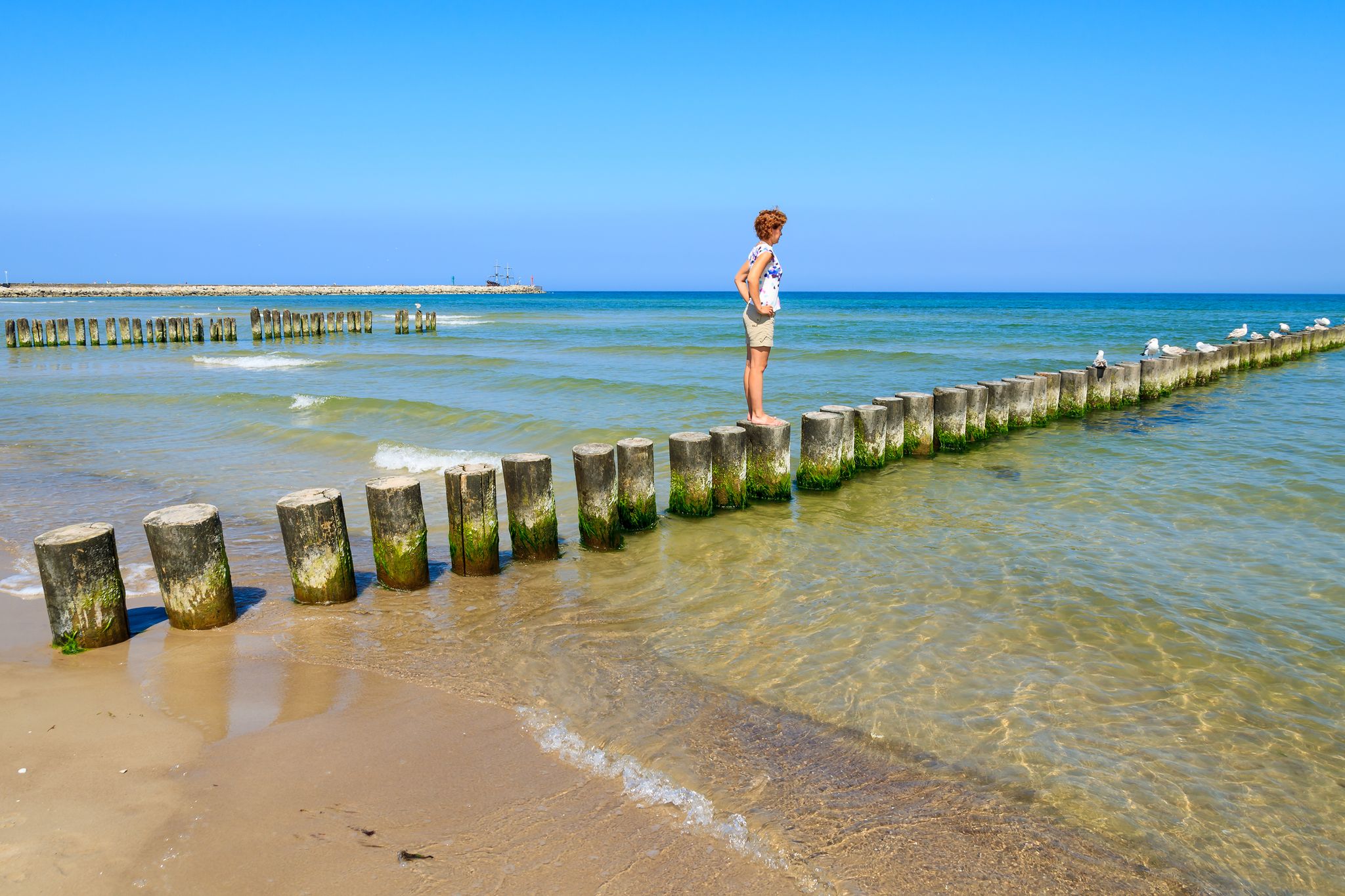 Young woman tourist standing on wooden breakwaters on beach in Ustka seaside town, Baltic Sea, Poland