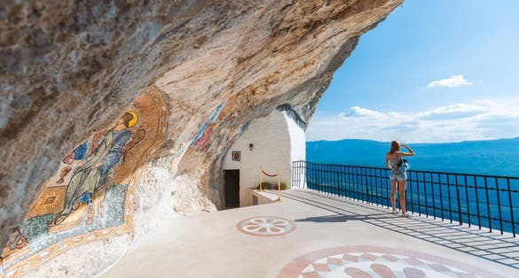 Photo of girl on viewpoint of Monastery Ostrog of the Serbian Orthodox Church situated against an almost vertical mountain with blue cloudy sky and mountains in background, Montenegro.