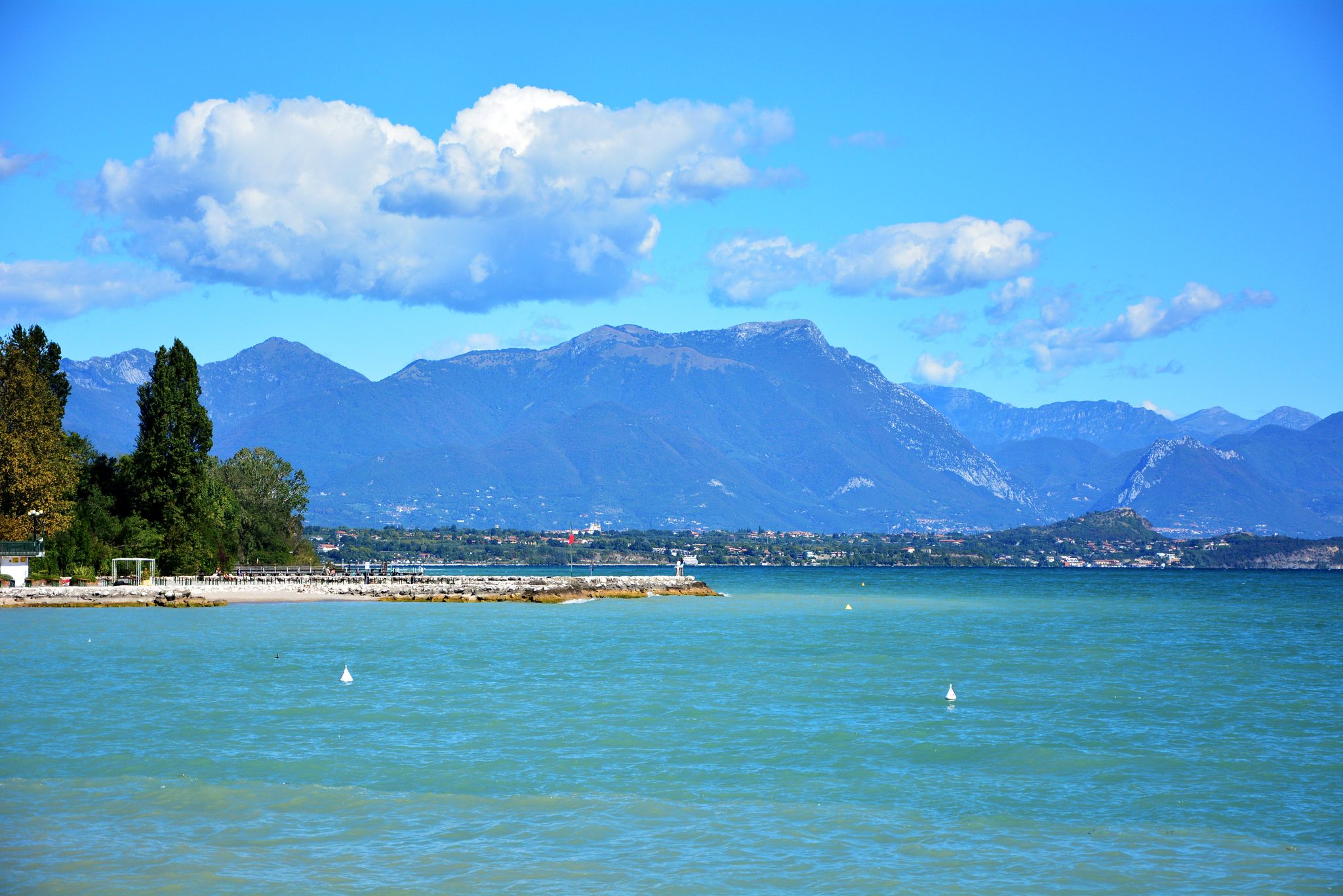 Photo of Old harbour Porto Vecchio with motor boats on turquoise water, green trees and traditional buildings in historical centre of Desenzano del Garda town, Northern Italy.