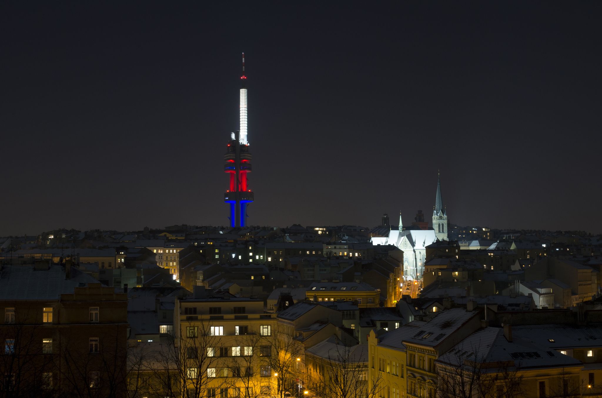 Photo of tall Zizkov television tower, old church and roofs of townhouses. Buildings are lit during night, in Prague, Czech Republic.