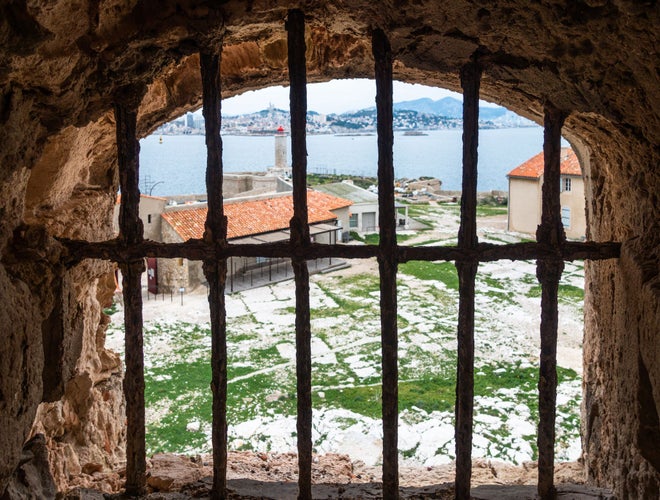 View through prison bars at Château d-If in Marseille, France, the historic island fortress made famous by The Count of Monte Cristo, overlooking the sea, lighthouse, and red-roofed buildings..jpg