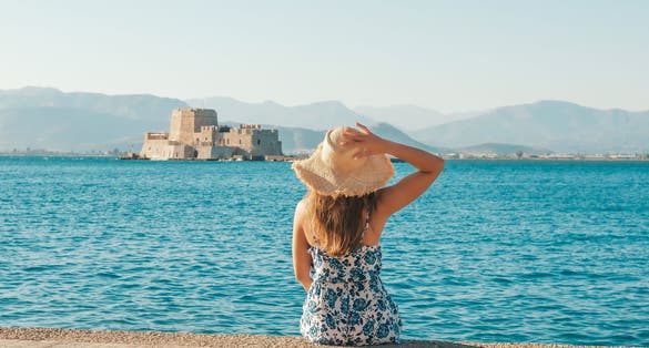 Photo of happy tourist sitting on the beach looking at the Bourtzi water castle is a small island with a fortress at the coast of Nafplio in Greece.
