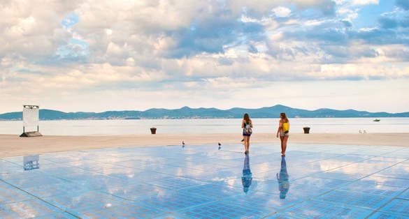 Photo of two girls walking on top of Sun Salutation installation near the sea shore against the background of a hill range and cloudy morning sky, Zadar, Croatia.