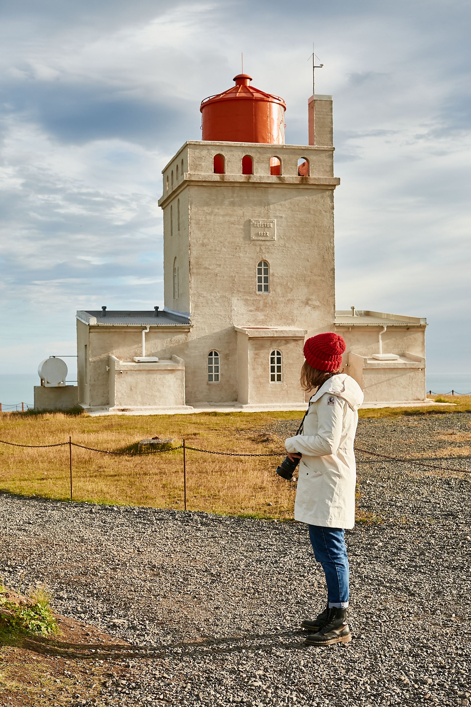 photo of young female tourist with photo camera looking at dyrholaey lighthouse located on the top of the cliff in Iceland near vik I myrdal village.