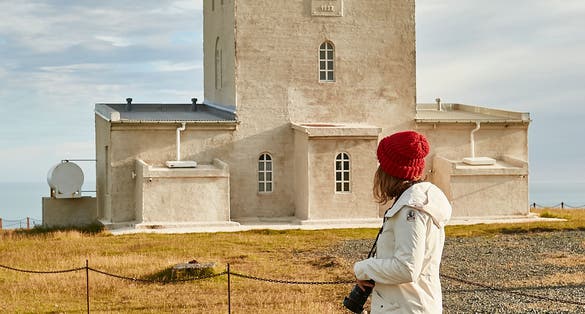 photo of young female tourist with photo camera looking at dyrholaey lighthouse located on the top of the cliff in Iceland near vik I myrdal village.