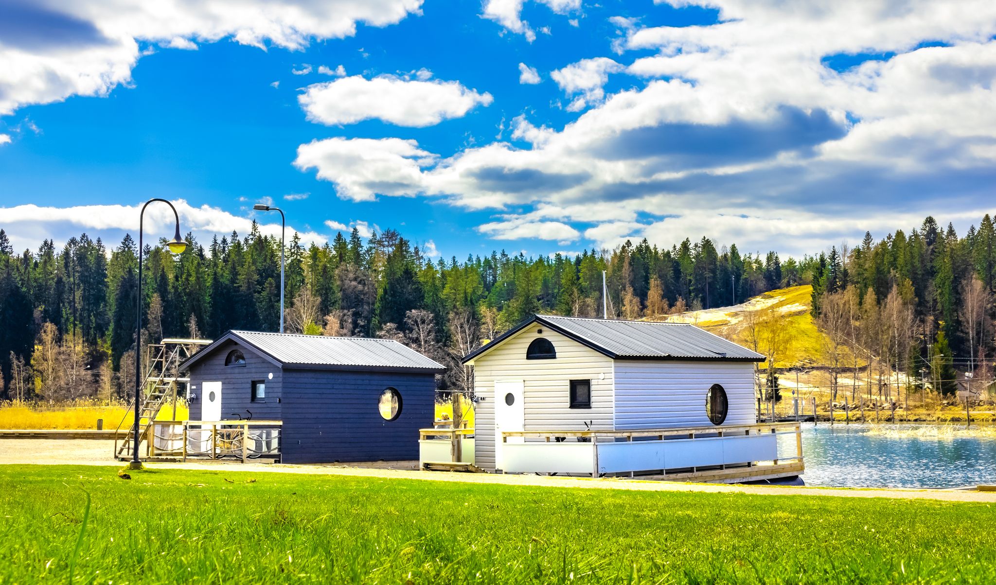 New modern cottages on water in Porvoo, Finland. Two small wooden houses under blue sky with white clouds at sunny day. Bright green lawn in front, pine trees forest behind. Cabin to relax in nature.