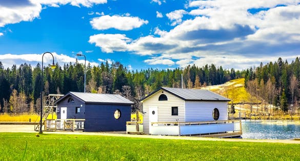 New modern cottages on water in Porvoo, Finland. Two small wooden houses under blue sky with white clouds at sunny day. Bright green lawn in front, pine trees forest behind. Cabin to relax in nature.