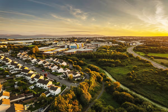 An aerial view of Galway, Ireland, at sunset, showing houses, greenery, roads, and the distant coastline..png