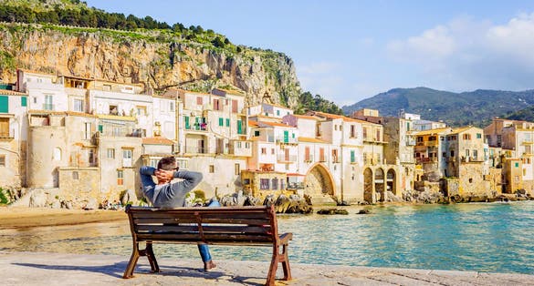 Photo of Young man sitting on bench, enjoying view of Cefalu coastline on Sicily island, Italy.
