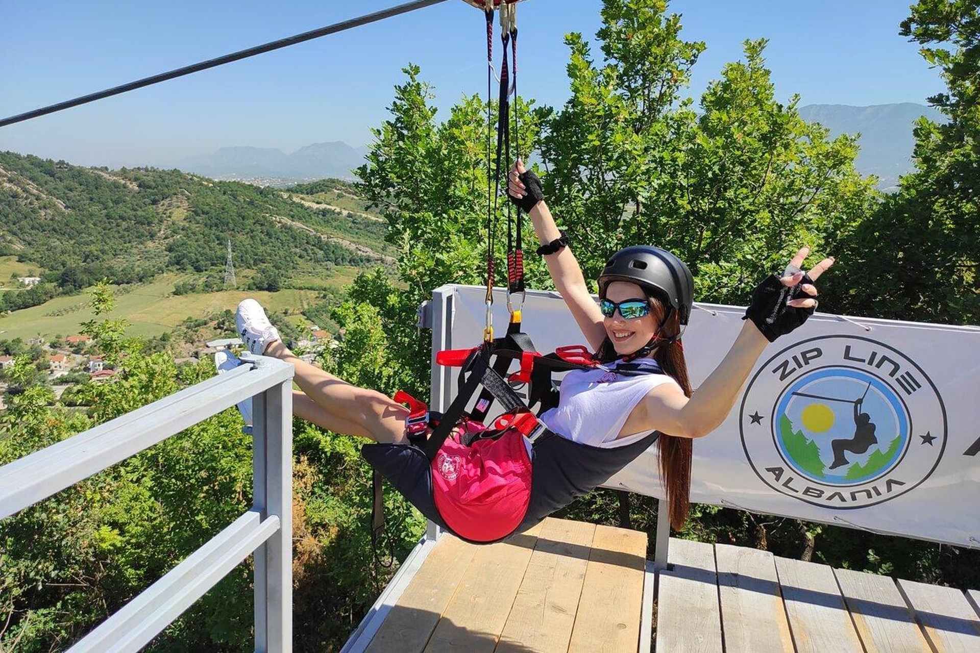 A smiling woman in zip-lining gear gets ready to ride at Zip Line Albania in Petrela, near Tirana, with green hills behind her..jpg