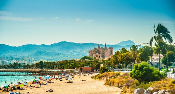 Photo of the beautiful beach of Palma de Mallorca with people lying on sand and the gorgeous cathedral building visible in background.