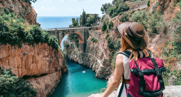 A girl in a cowboy hat with a backpack enjoys an incredibly beautiful view of a stone bridge over the gorge Fiordo di Furore.