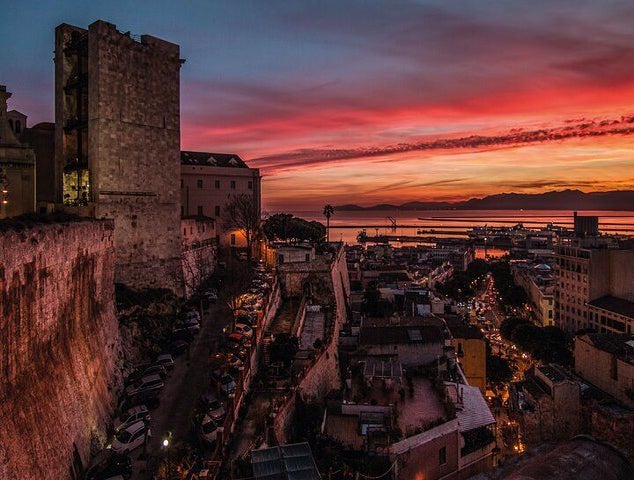 A historic cityscape at sunset with ancient stone buildings, the Torre dell’Elefante, a bustling street, and a harbor reflecting the colorful sky..jpg