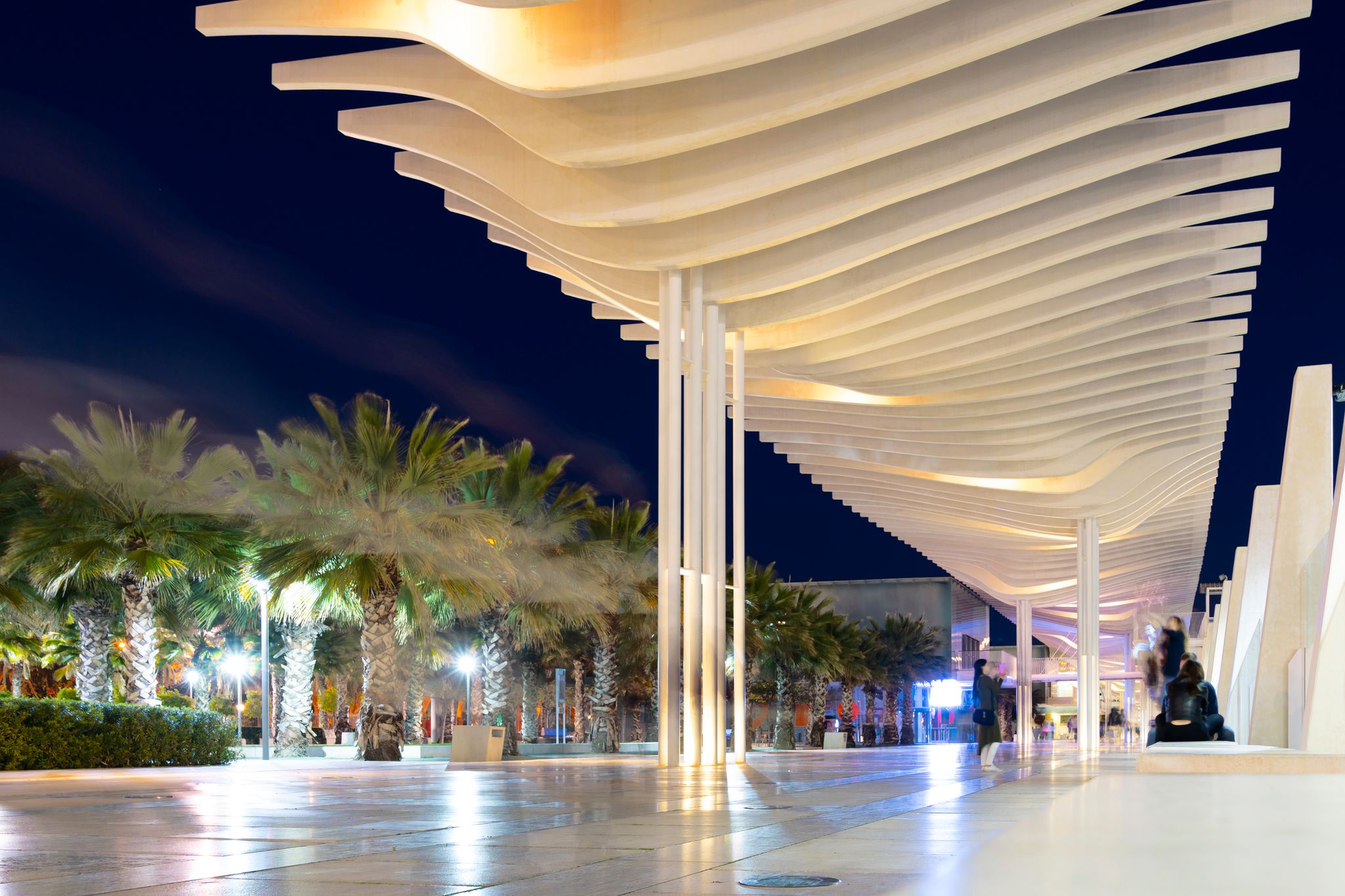 Photo of Great view of Malaga city in the night: promenade of Palmeral de las Sorpresas, beautiful white wawe of Pergola. Happy young people in a modern port area of Malaga, Andalusia, southern Spain, Europa.