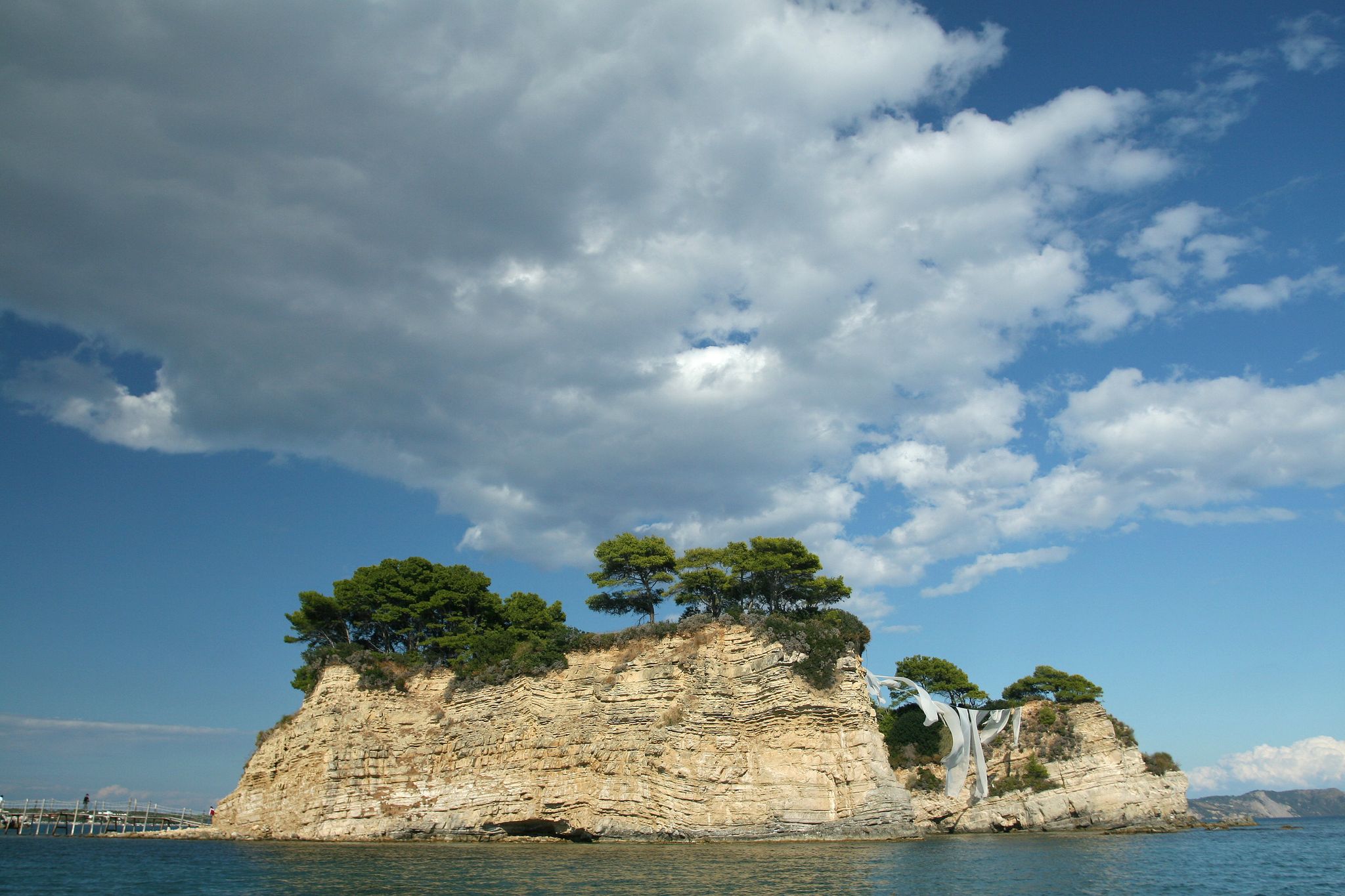 photo of view of Cameo Island, the most picturesque place in Laganas Bay, with pine trees growing in the rocks and white cloth blowing in the wind, Agios Sostis, Zakynthos, Greece,Laganas Greece.