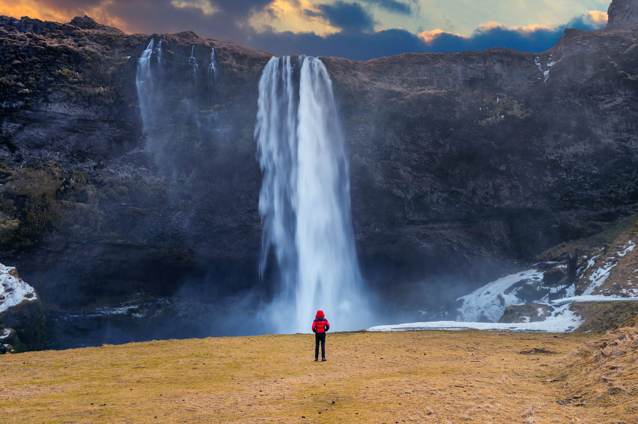 photo of seljalandsfoss waterfall in Iceland. Guy in red jacket looks at seljalandsfoss waterfall.
