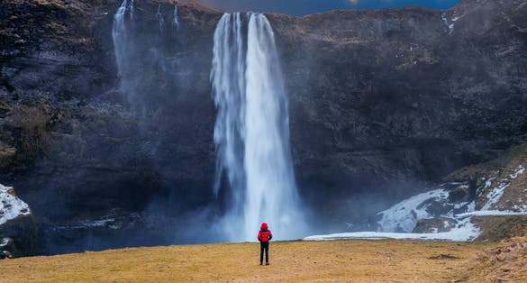 photo of seljalandsfoss waterfall in Iceland. Guy in red jacket looks at seljalandsfoss waterfall.