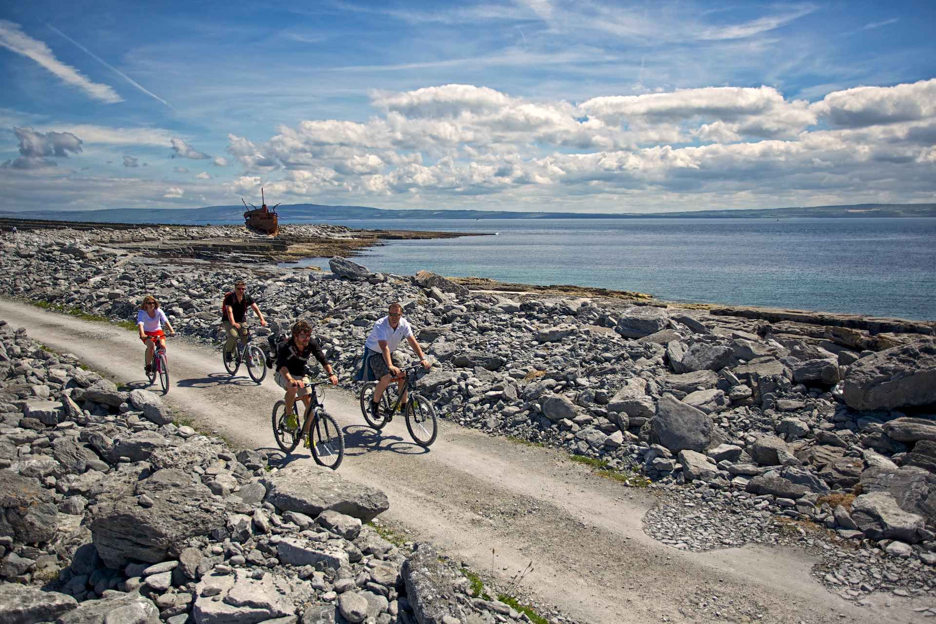 Four people ride bicycles on a rocky coastal path on the Aran Islands, with a rusted shipwreck in the background..jpg