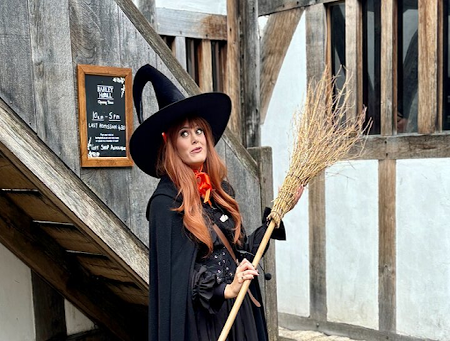 A woman dressed as a witch with a black hat and cloak holds a broomstick in front of a rustic wooden and stone building..png