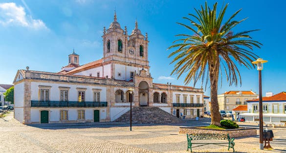 Photo of Sanctuary of Our Lady of Nazare catholic church in cobblestone square with palm trees in Sitio hilltop da Nazare town, clear blue sky on sunny summer day, Leiria District, Oeste region, Portugal.