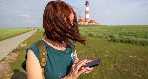 photo of view of A woman uses her mobile phone in front of the Westerheversand lighthouse, Westerheversand, Germany.