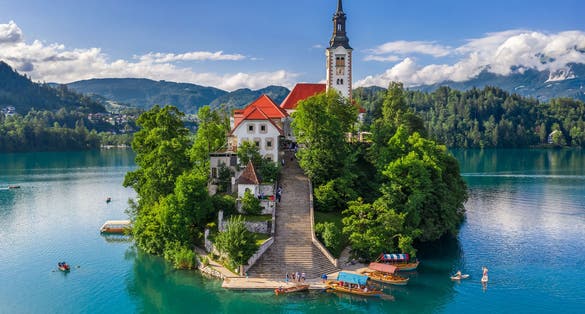 Photo of Bled, Slovenia - Aerial view of beautiful Pilgrimage Church of the Assumption of Maria on a small island at Lake Bled (Blejsko Jezero) and lots of Pletna boats on the lake at summer time with blue sky.