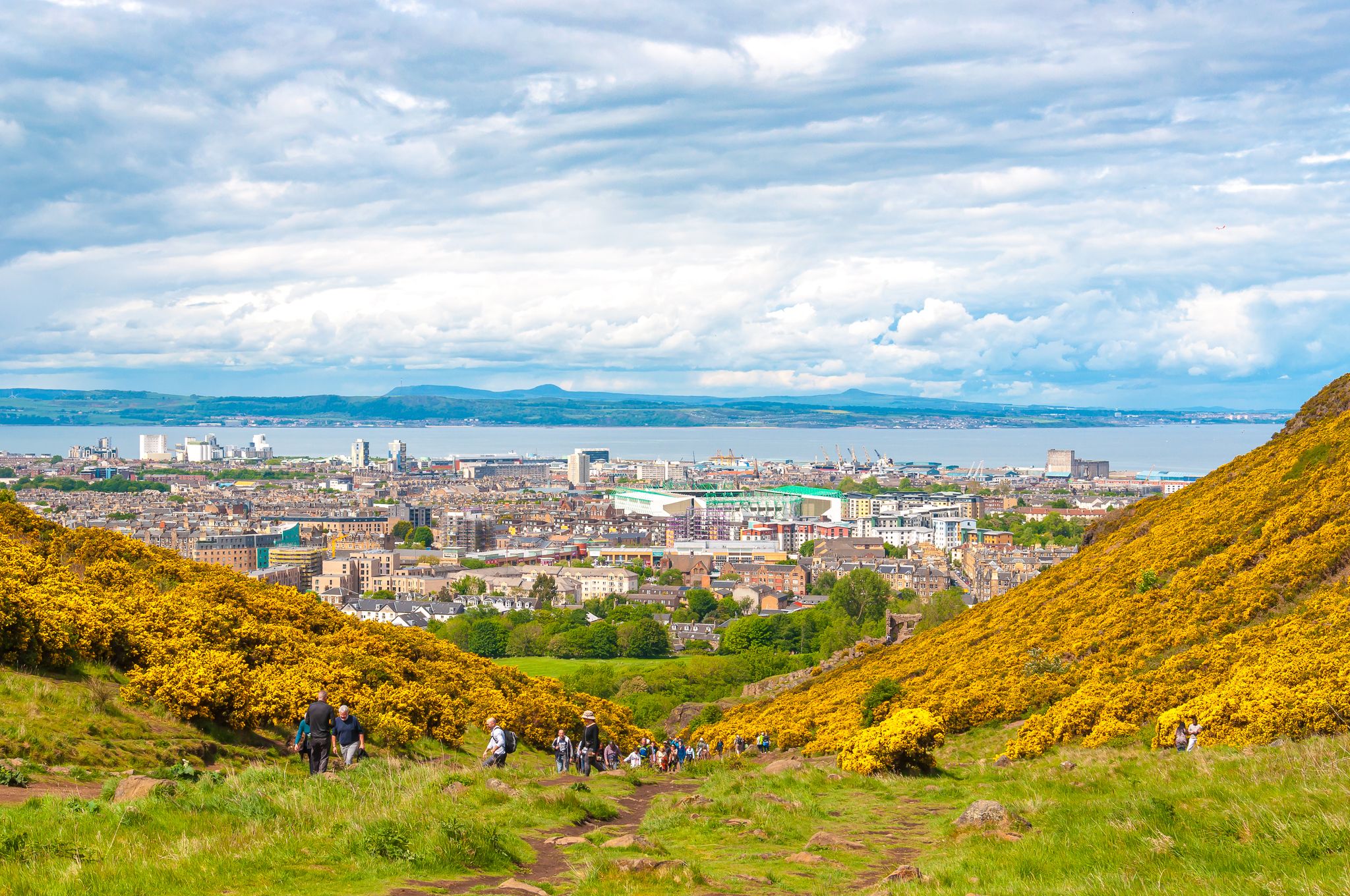 Unrecognizable people in Holyrood Park surrounded by yellow flowers with panorama of Edinburgh background. Concept: Scottish urban landscapes