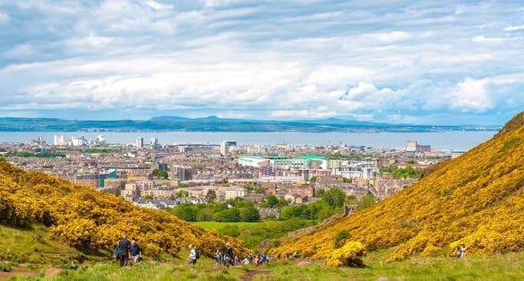 Unrecognizable people in Holyrood Park surrounded by yellow flowers with panorama of Edinburgh background. Concept: Scottish urban landscapes