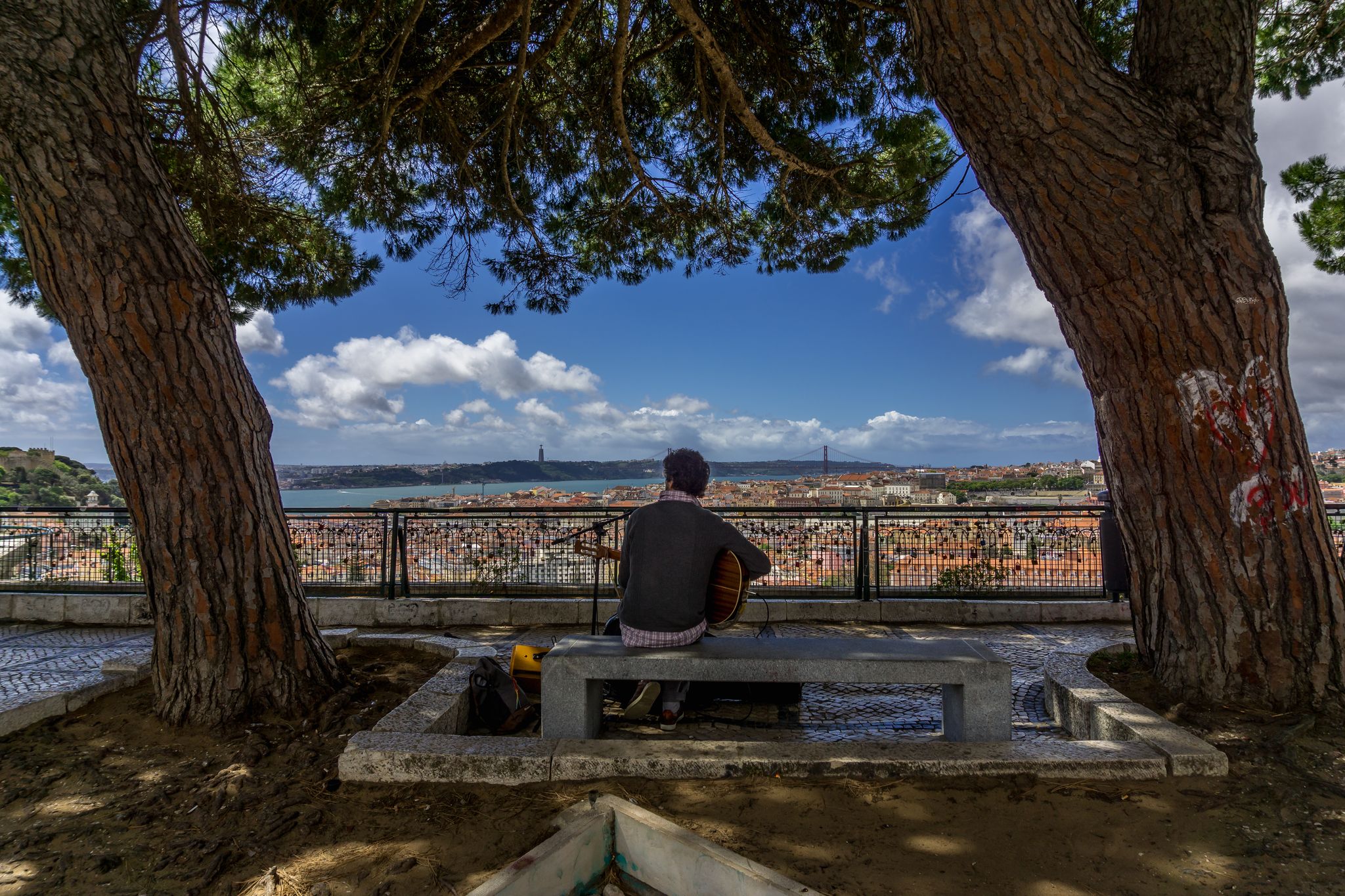 Photo of beautiful shot of a man sitting on a bench and playing the guitar. Miradouro da Nossa Senhora do Monte viewpoint of Lisbon, Portugal.