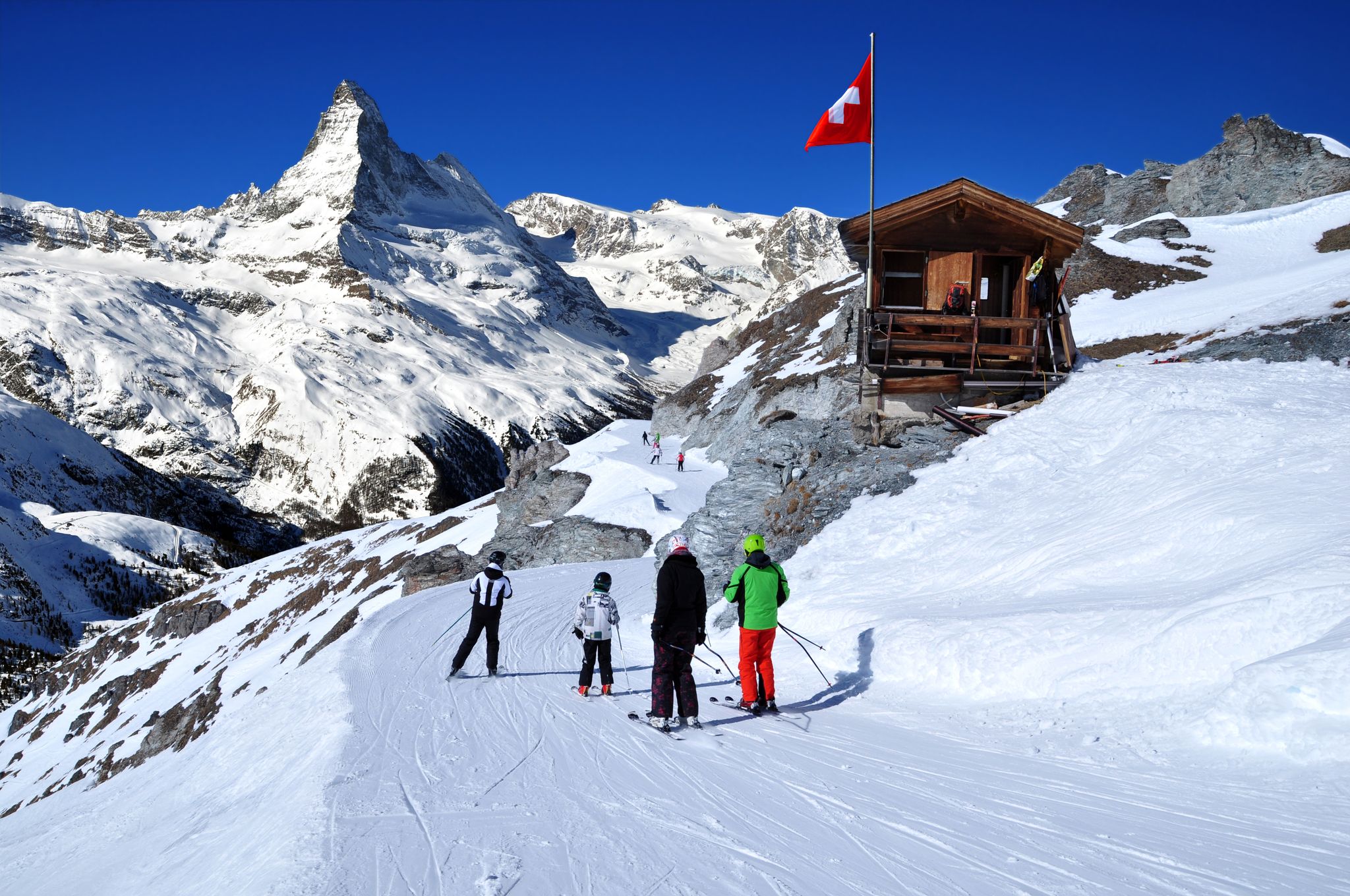 photo of skiers on the way to ski slope in Swiss Alps in sunny day, Zermatt, Matterhorn behind. Small wooden house with red swiss flag near by ski slope. Mountains and skiing resort photo manipulation, Switzerland.