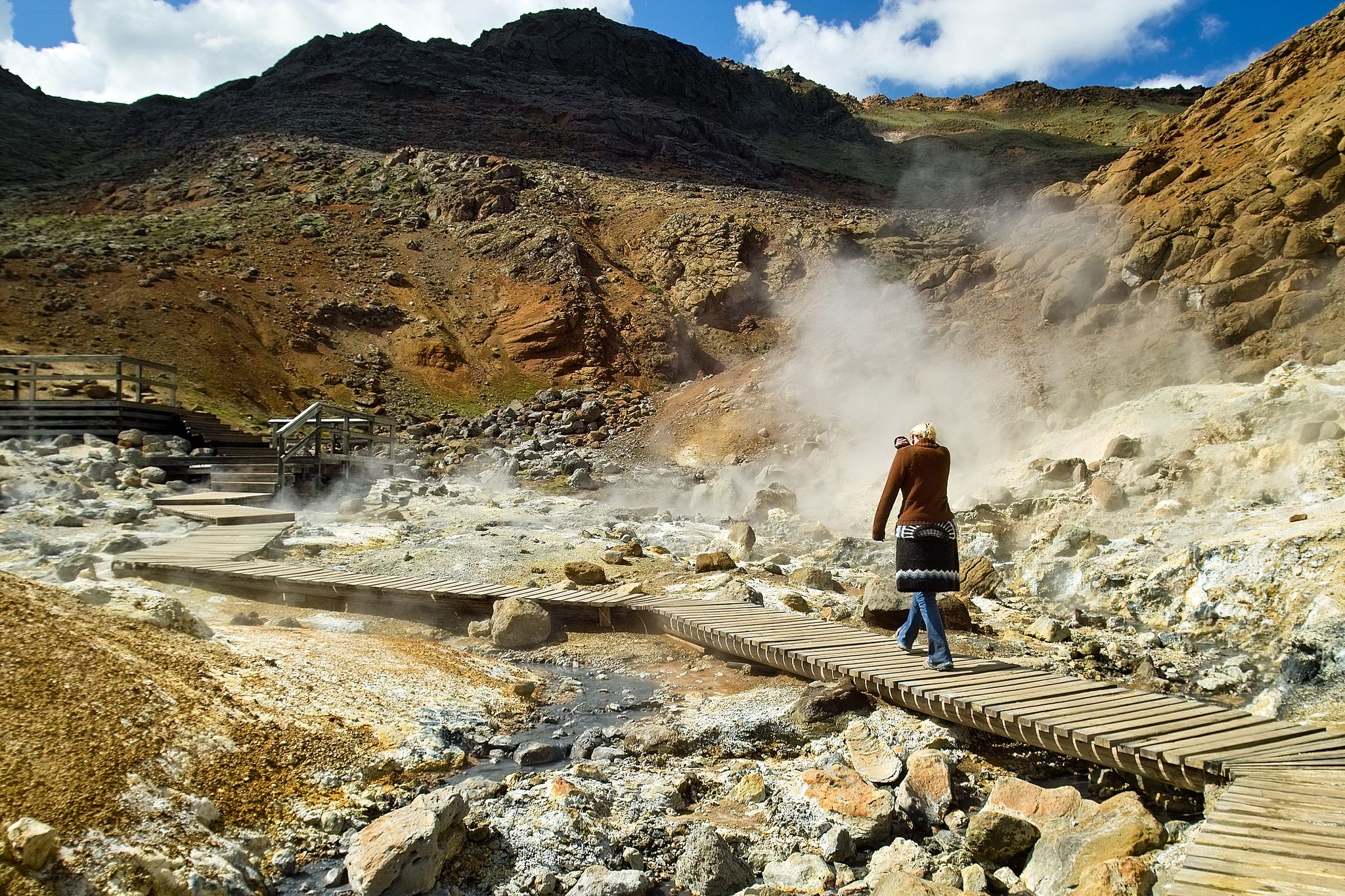 photo of woman walking on wooden pathway trying to breath through a sweater sleeve to avoid sulfur smell - in srýsuvík / seltún geothermal area in Reykjanes peninsula in Iceland.