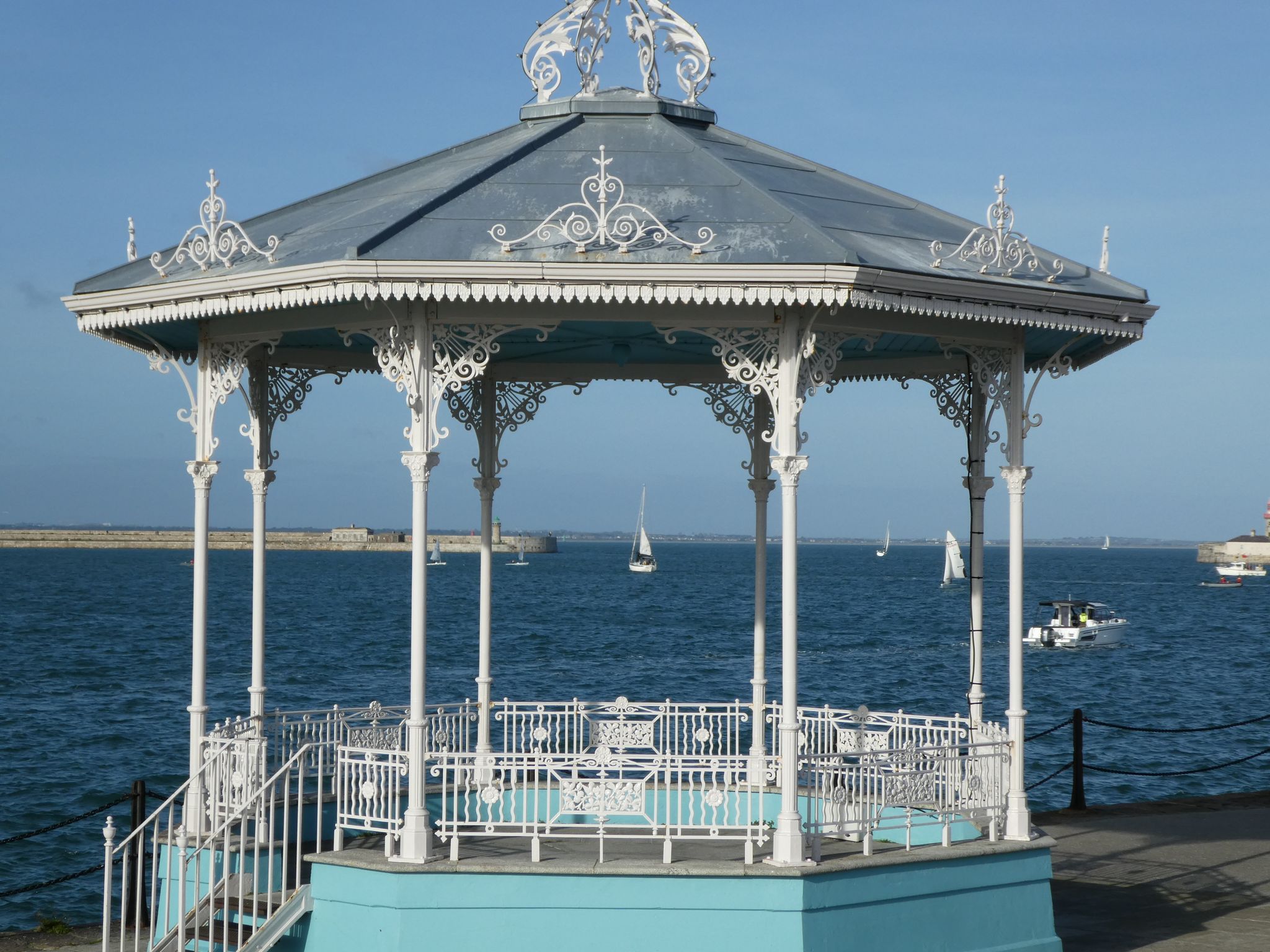 photo of view of A pleasure yacht can be seen framed by the old white iron work of the band stand at Dun Laoghaire against a bright blue sky and deep blue water, Dún Laoghaire, Ireland.