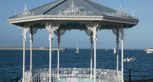 photo of view of A pleasure yacht can be seen framed by the old white iron work of the band stand at Dun Laoghaire against a bright blue sky and deep blue water, Dún Laoghaire, Ireland.