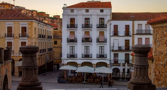 Photo of A view of a section of Plaza Mayor (main square) featuring white buildings, stone buildings, outdoor seating at a restaurant and stone columns in the later afternoon. Cáceres, Extramadura, Spain.