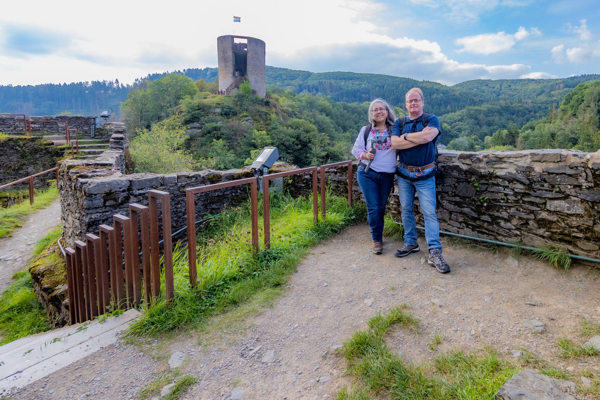 photo of mature tourist couple smiling and looking at camera, Esch-sur-Sûre castle ruins, tower with waving flag, countryside with hills with abundant green trees in the background, cloudy day in Luxembourg.