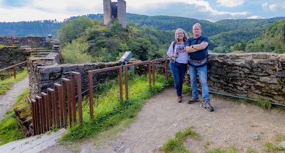 photo of mature tourist couple smiling and looking at camera, Esch-sur-Sûre castle ruins, tower with waving flag, countryside with hills with abundant green trees in the background, cloudy day in Luxembourg.