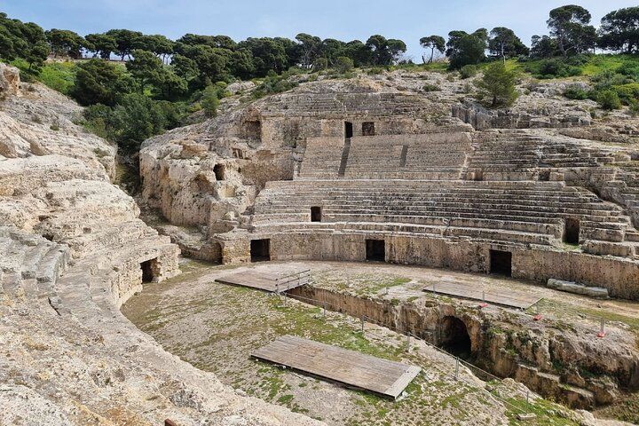 An ancient stone amphitheater with tiered seating, arched entrances, and a stage, surrounded by trees and hills..jpg
