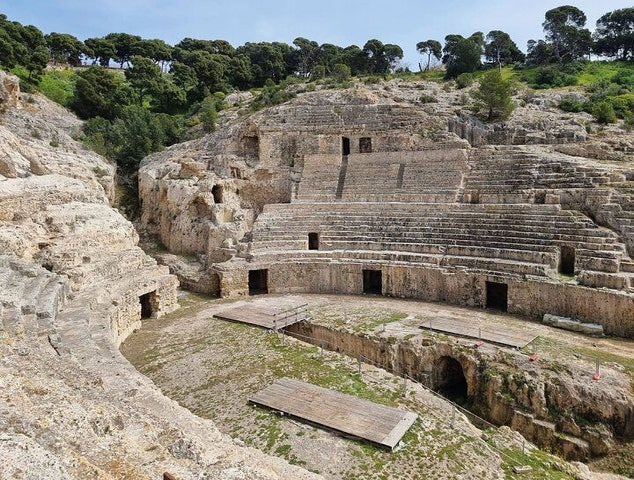 An ancient stone amphitheater with tiered seating, arched entrances, and a stage, surrounded by trees and hills..jpg