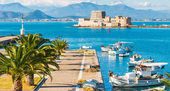Photo of beautiful port of Nafplio city in Greece with small boats, palm trees and Bourtzi castle on the water.